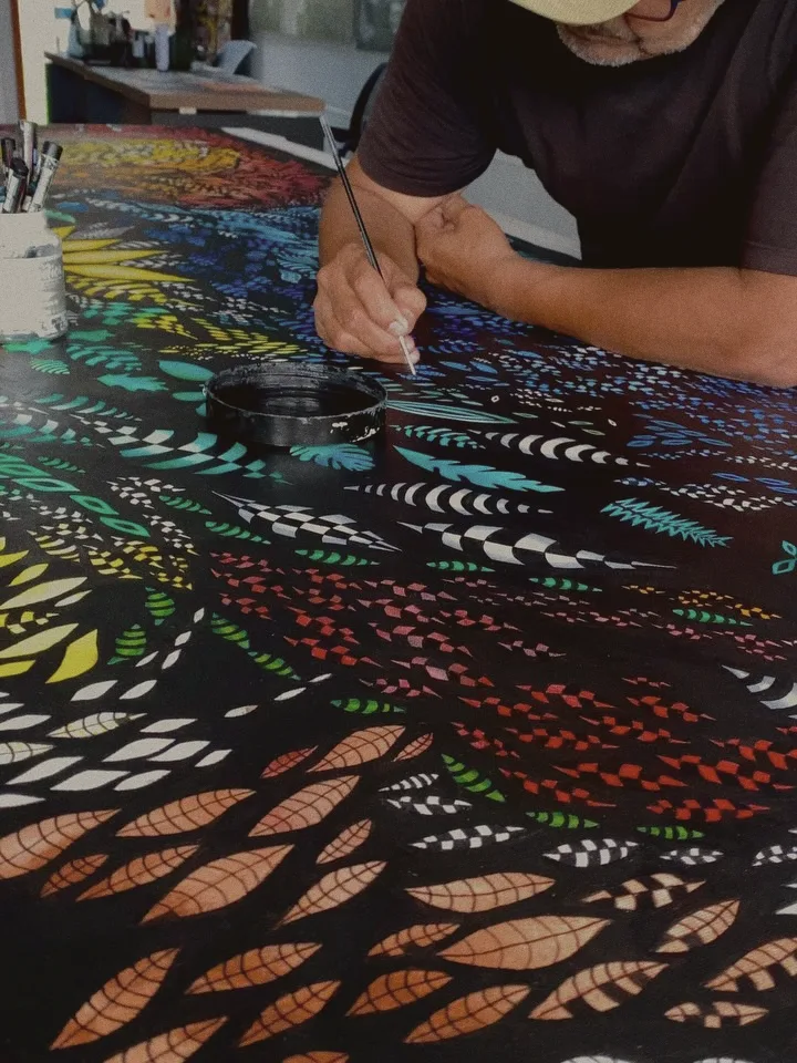 Almir pintando à mão, com pincel fino, folhas coloridas sobre fundo preto